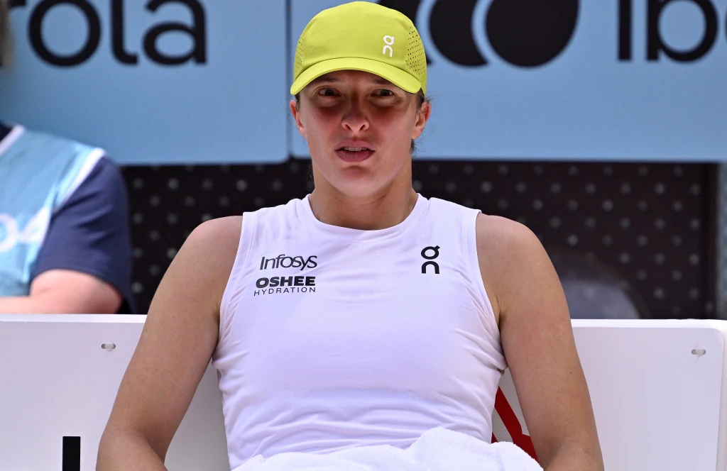 Iga Świątek Sportowiec w białej koszulce i żółtej czapce siedzi podczas przerwy na tle reklamy turnieju tenisowego.