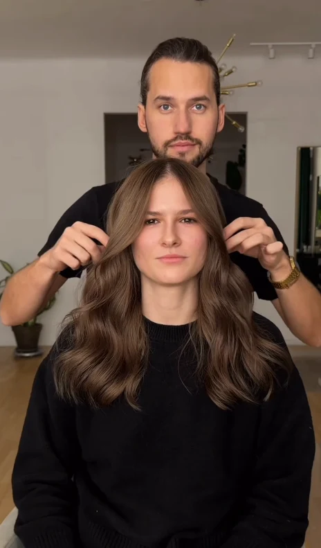 Mężczyzna stojący za kobietą, która siedzi na krześle, układa jej rozpuszczone brązowe włosy. Oboje mają spokojne wyraz twarzy. W tle widać jasne, minimalistyczne wnętrze.