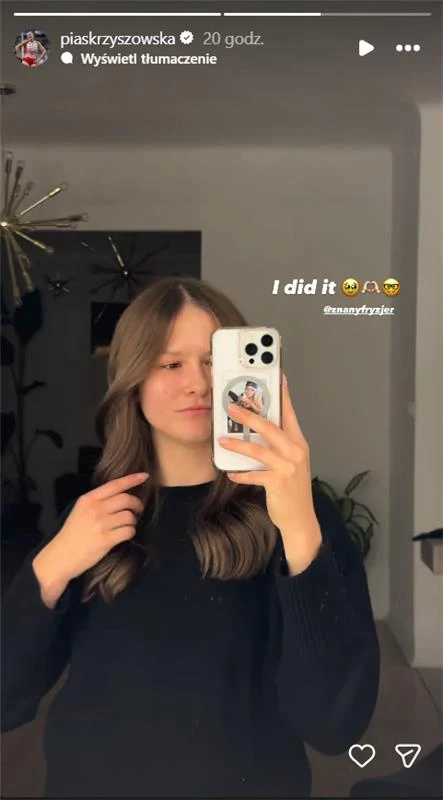 Młoda kobieta z długimi, falowanymi włosami robi selfie w lustrze, trzymając telefon z białym etui, ubrana w czarny sweter, w tle nowoczesne wnętrze z rośliną doniczkową oraz złotą lampą.