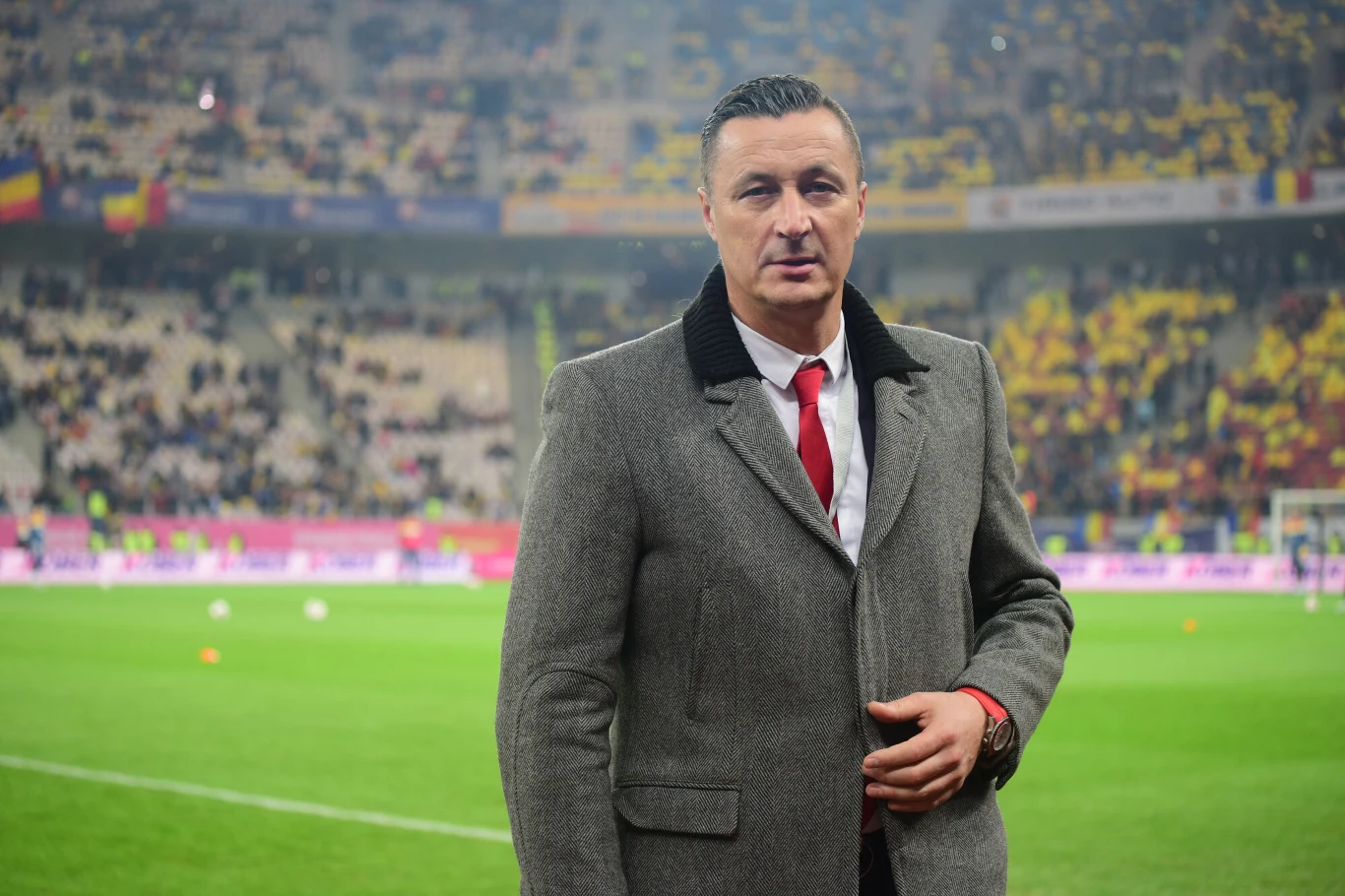 Mężczyzna w eleganckim płaszczu i zawiązanym czerwonym krawatem stoi na murawie stadionu piłkarskiego, za nim tłum kibiców na trybunach wypełnionych żółtymi flagami i transparentami.