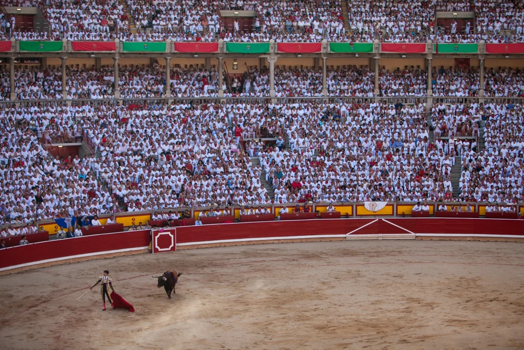 Arena korridy wypełniona tłumem widzów ubranych na biało, matador konfrontuje się z bykiem na środku areny.