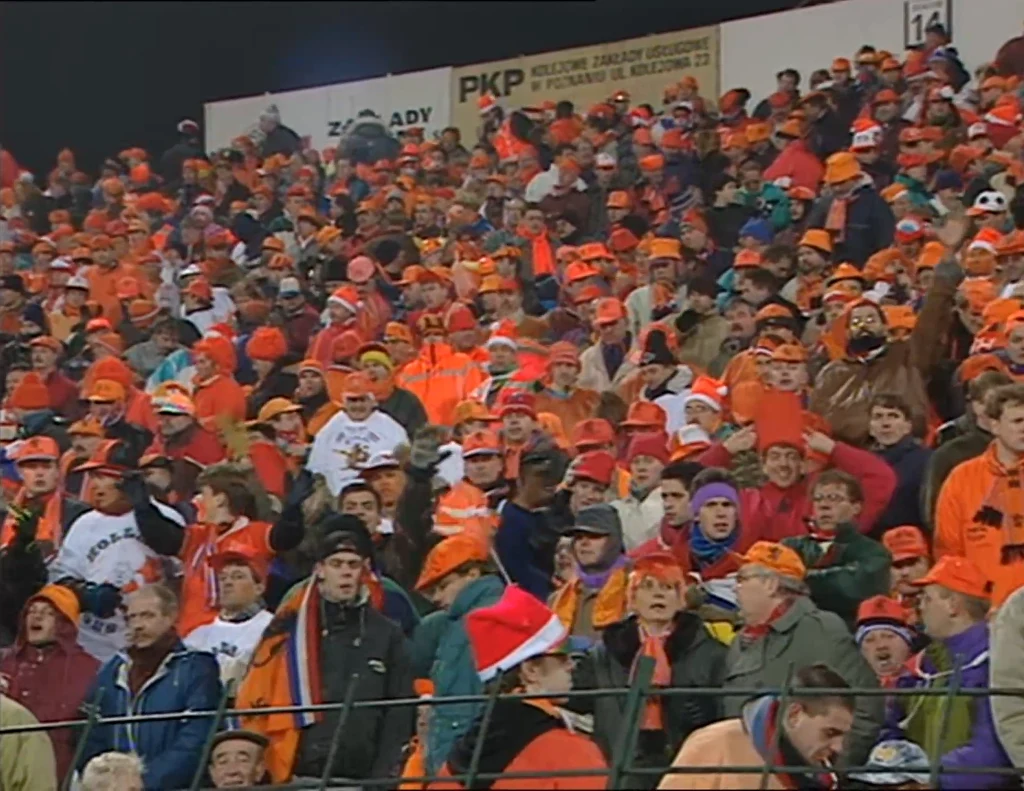Inwazja holenderskich kibiców na Poznań przy okazji meczu Polska - Holandia Tłum kibiców ubranych w pomarańczowe czapki, szaliki i ubrania, zgromadzony na trybunach stadionu. W tle widoczne są banery reklamowe. Atmosfera jest radosna i pełna energii.