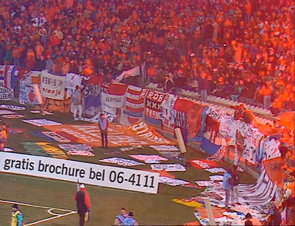 Holenderscy kibice podczas meczu Polska - Holandia Tłum kibiców na stadionie, ubranych w pomarańczowe stroje, trzymający i rozwieszający liczne flagi oraz banery na ogrodzeniu, z wyrazistym, czerwonym oświetleniem i reklamą z numerem telefonu na pierwszym planie.