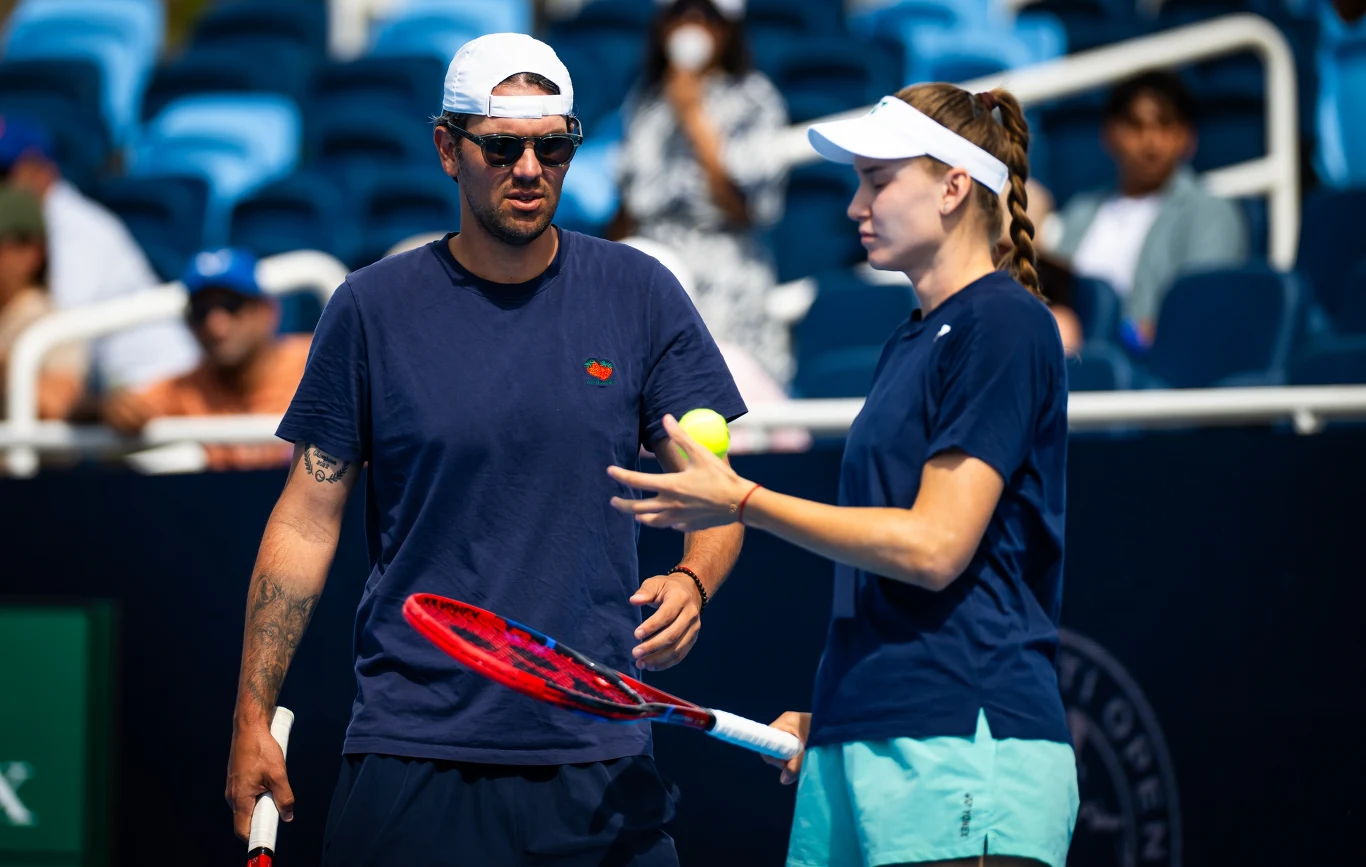 Dwójka osób na korcie tenisowym podczas rozmowy, jedna z osób trzyma rakietę tenisową i piłkę, tło stanowią niebieskie trybuny oraz kilku widzów.