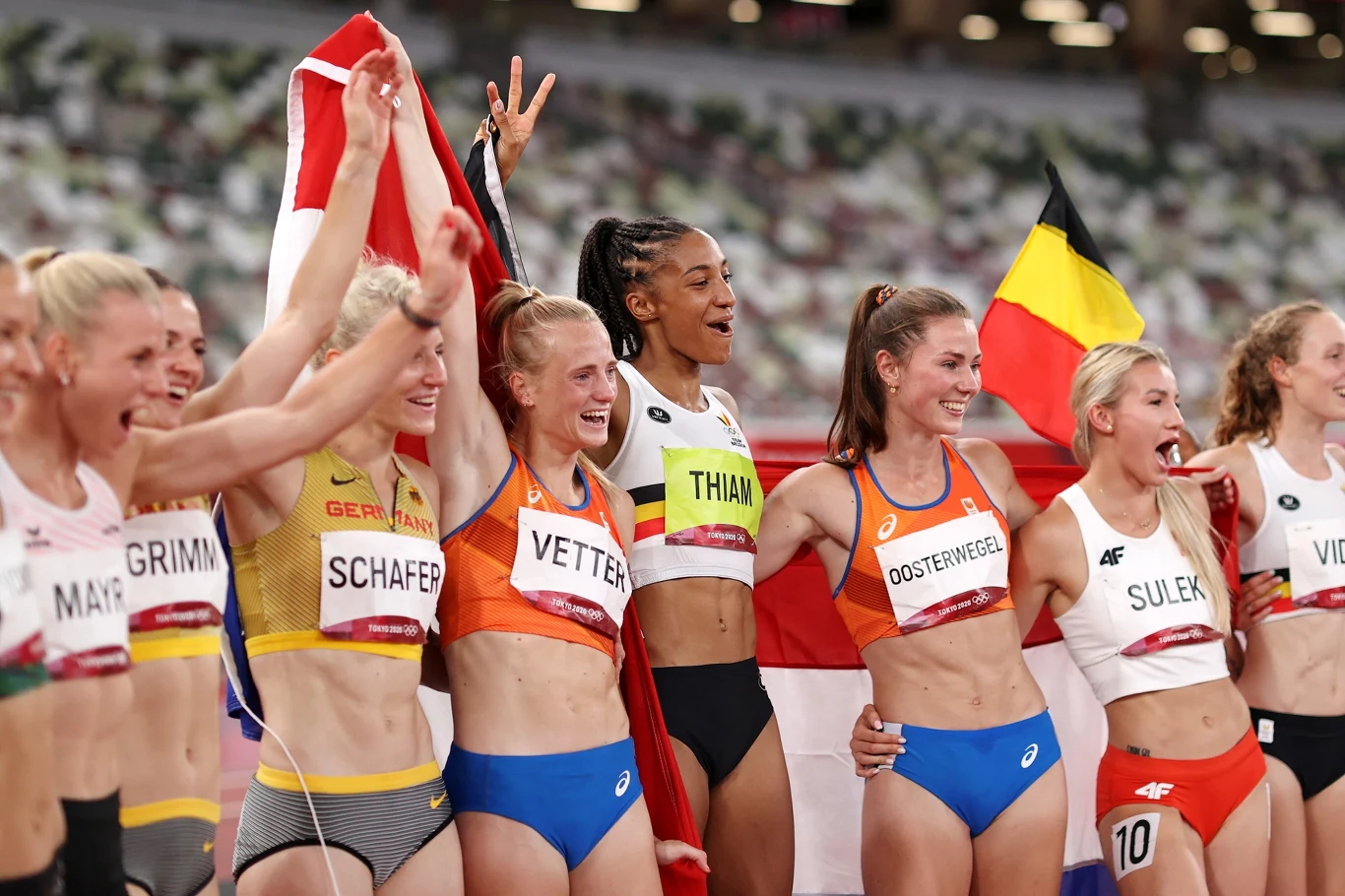 Grupa radosnych lekkoatletek w strojach sportowych po zakończeniu zawodów, trzymają flagi narodowe i świętują wspólnie zdobyte sukcesy, część z nich obejmuje się i uśmiecha.