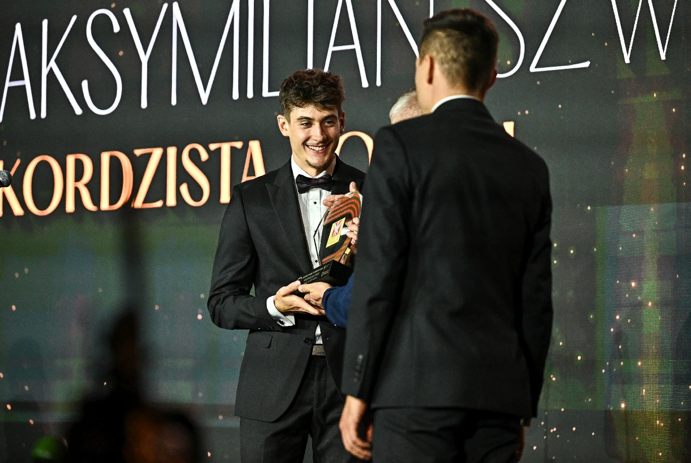 Młody mężczyzna w smokingu odbiera nagrodę trzymaną w obu dłoniach, uśmiecha się szeroko, obok stoją dwie osoby, jedno z nich widoczne od tyłu. Tło stanowi scena z dużym napisem i światłami.