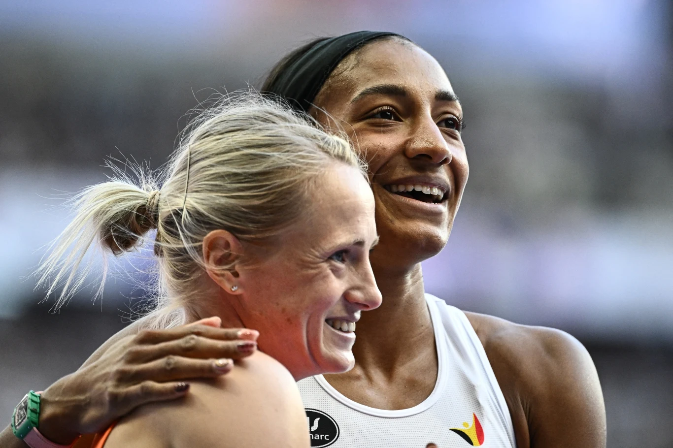 Dwie kobiety sportowczynie obejmują się i uśmiechają do siebie na tle stadionu, wyrażając radość po zakończonych zawodach lekkoatletycznych.
