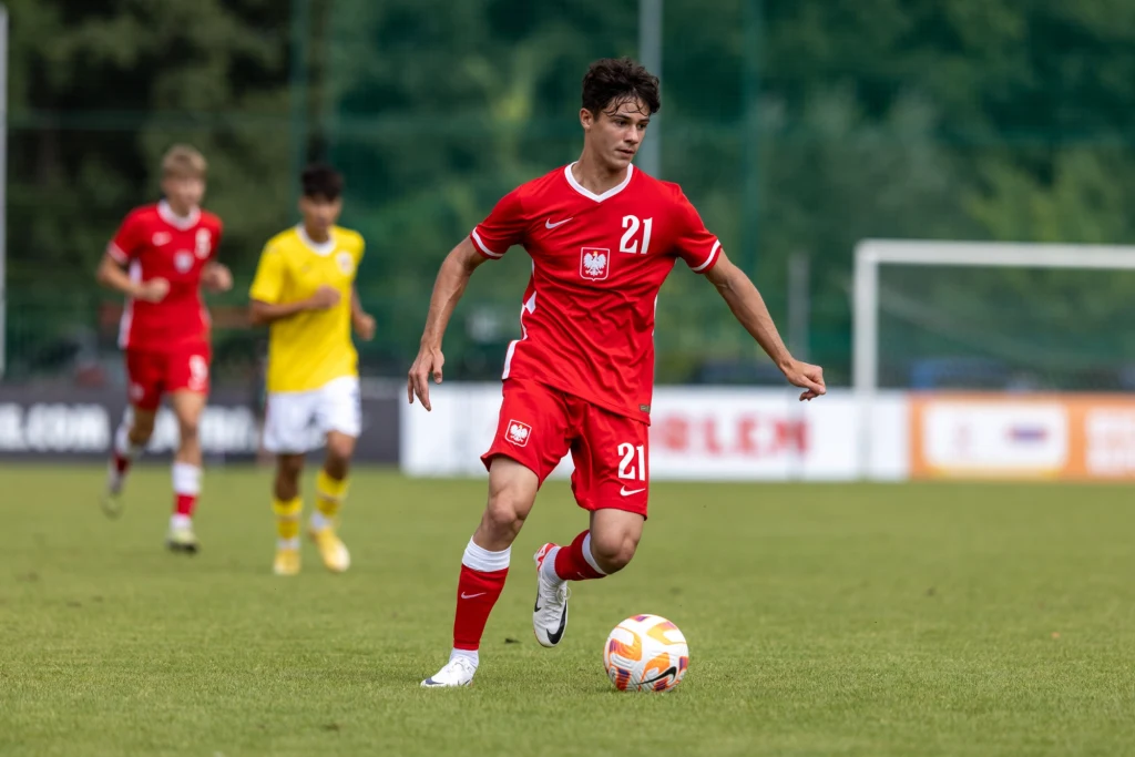 Piłkarz w czerwonym stroju z numerem 21 prowadzi piłkę po murawie podczas meczu, w tle widoczni są inni zawodnicy i bramkarz.