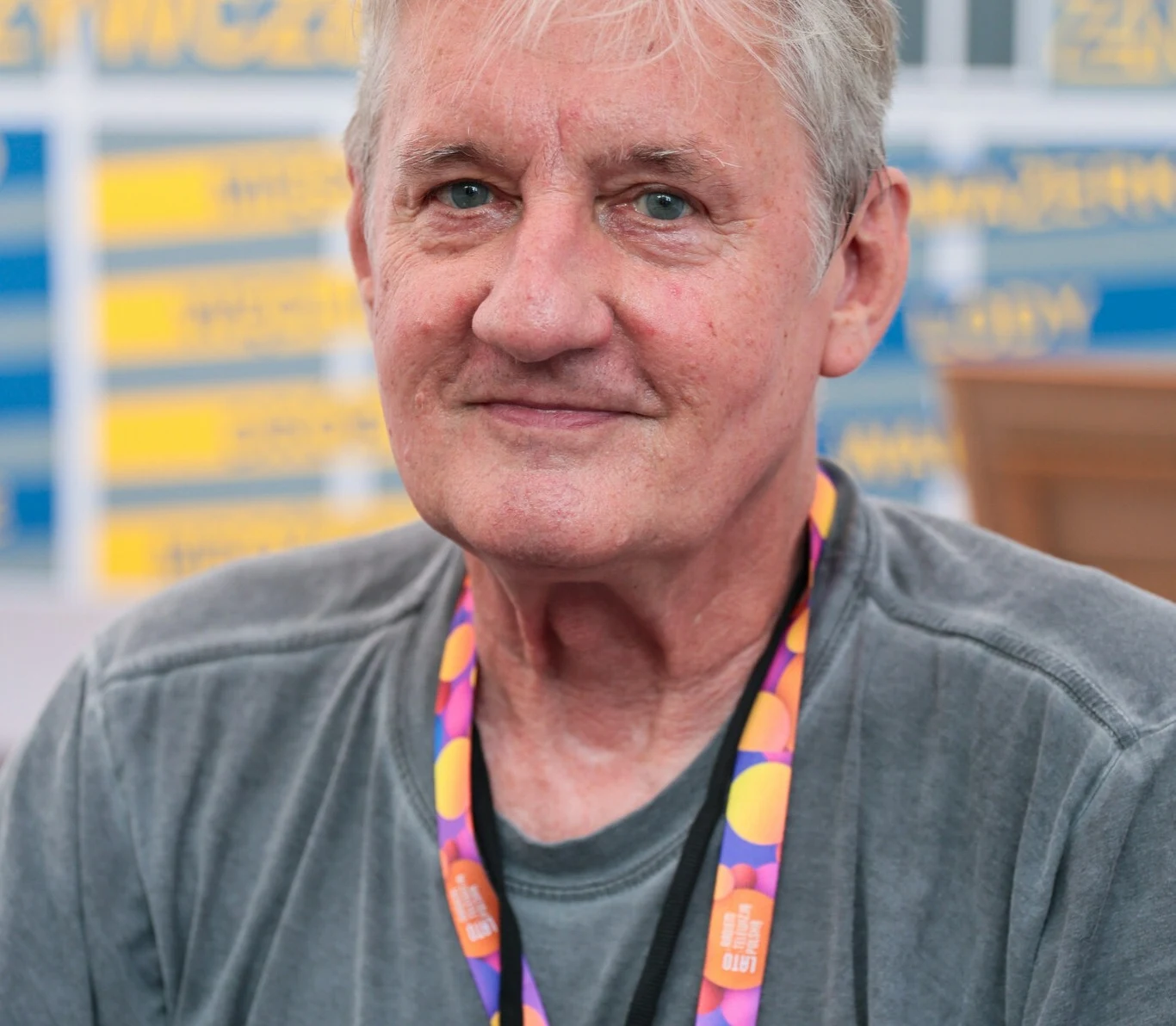Starszy mężczyzna z siwymi włosami siedzi uśmiechając się lekko do aparatu, ubrany w szarą koszulkę i kolorową smycz na szyi, za jego plecami widoczne jest rozmyte tło z żółto-niebieskimi tablicami.