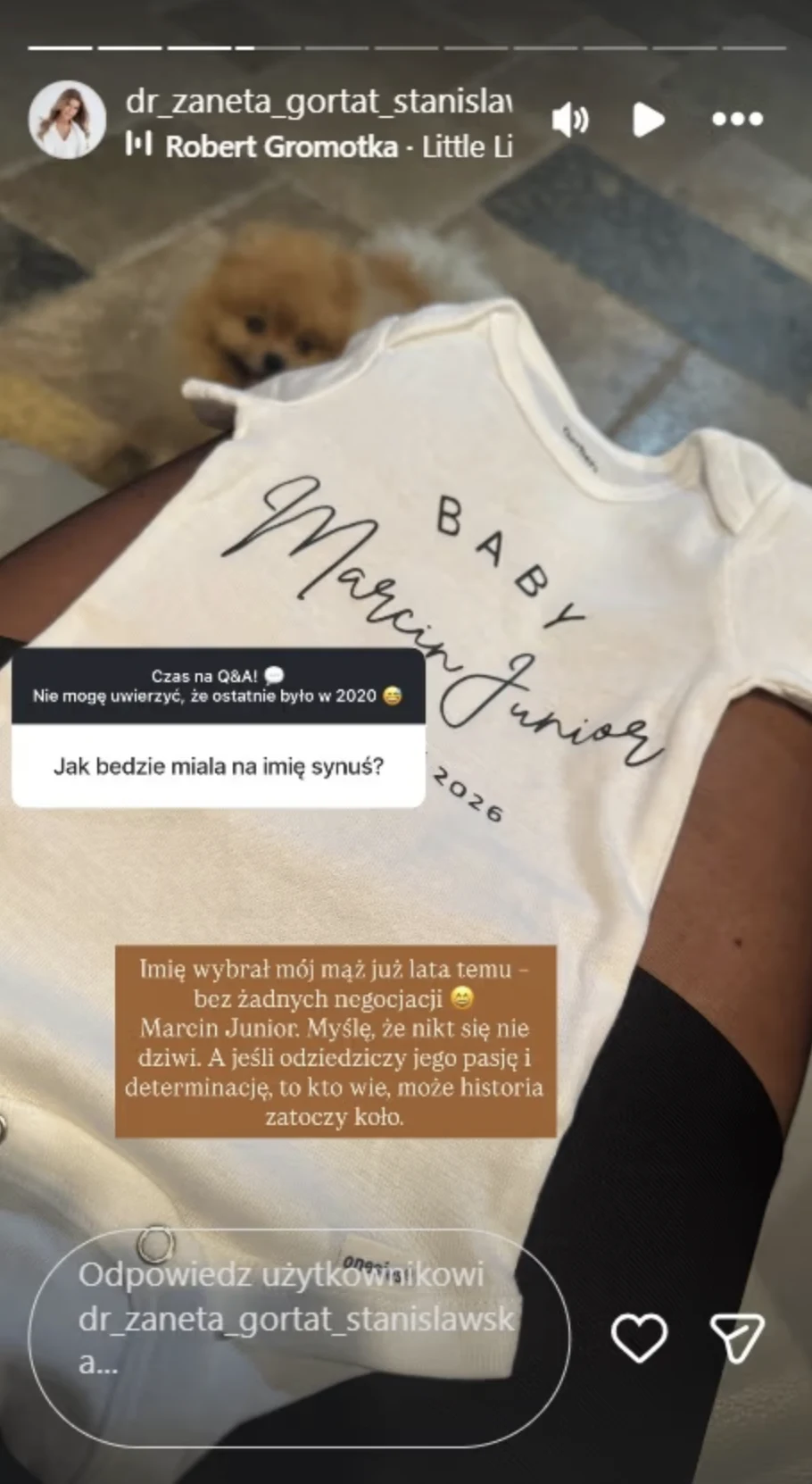 Białe body dziecięce z napisem 'Baby Marcin Junior 2026', na tle jasnego kocyka, w tle widoczny rozmazany pies. Na body nałożone są okienka z pytaniem i odpowiedzią w języku polskim dotyczące wyboru imienia dla syna.