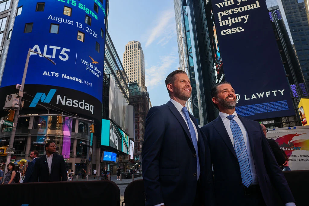 Dwóch elegancko ubranych mężczyzn w garniturach stoi w centrum Times Square na tle wieżowców i dużych ekranów LED, na których widoczne są logotypy Nasdaq oraz komunikaty reklamowe.