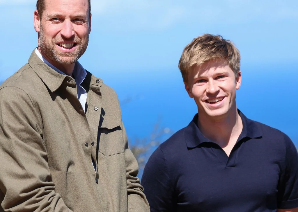 Książę William i Robert Irwin w 2024 roku Dwóch uśmiechniętych mężczyzn pozuje wspólnie na tle jasnoniebieskiego nieba i morza; jeden w jasnej kurtce, drugi w granatowej koszulce.
