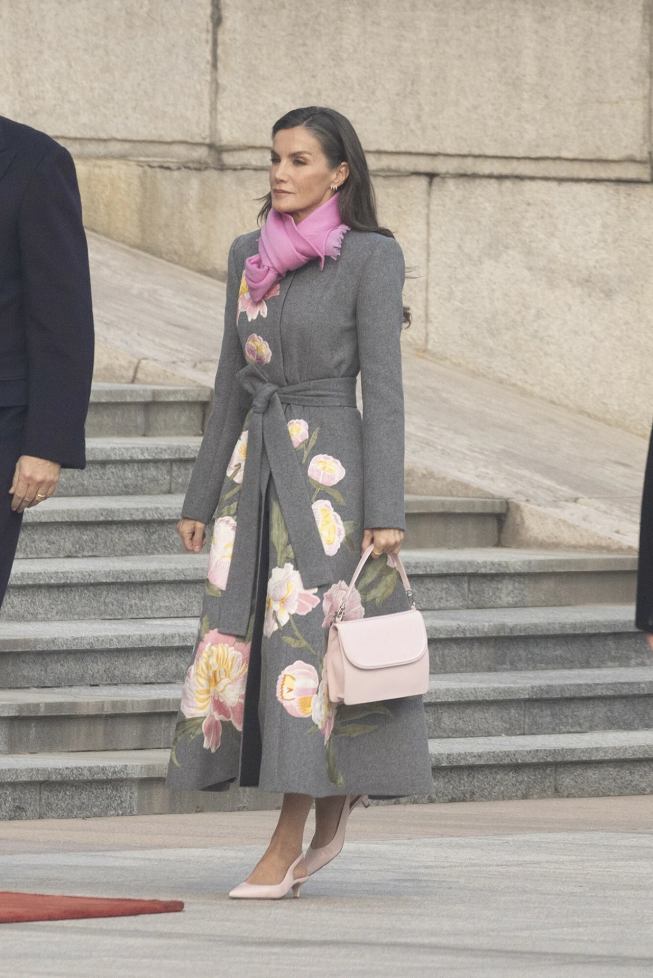 Elegancka kobieta ubrana w szary płaszcz z kwiatowymi zdobieniami, różowy szalik oraz jasnoróżowe buty na obcasie i torebkę, stojąca na schodach w miejskim otoczeniu.