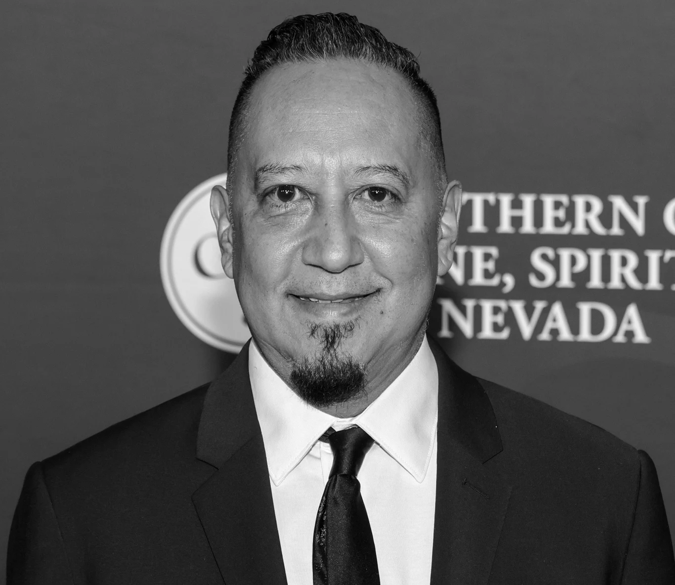 Cleto Escobedo III miał 59 lat Mężczyzna w średnim wieku w eleganckim garniturze z krawatem, stojący na tle ścianki z widocznymi napisami.