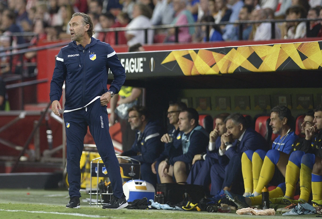 Trener drużyny piłkarskiej stojący przy linii bocznej podczas meczu, obok niego na ławce rezerwowych siedzą zawodnicy w niebiesko-żółtych strojach, w tle trybuny pełne kibiców.