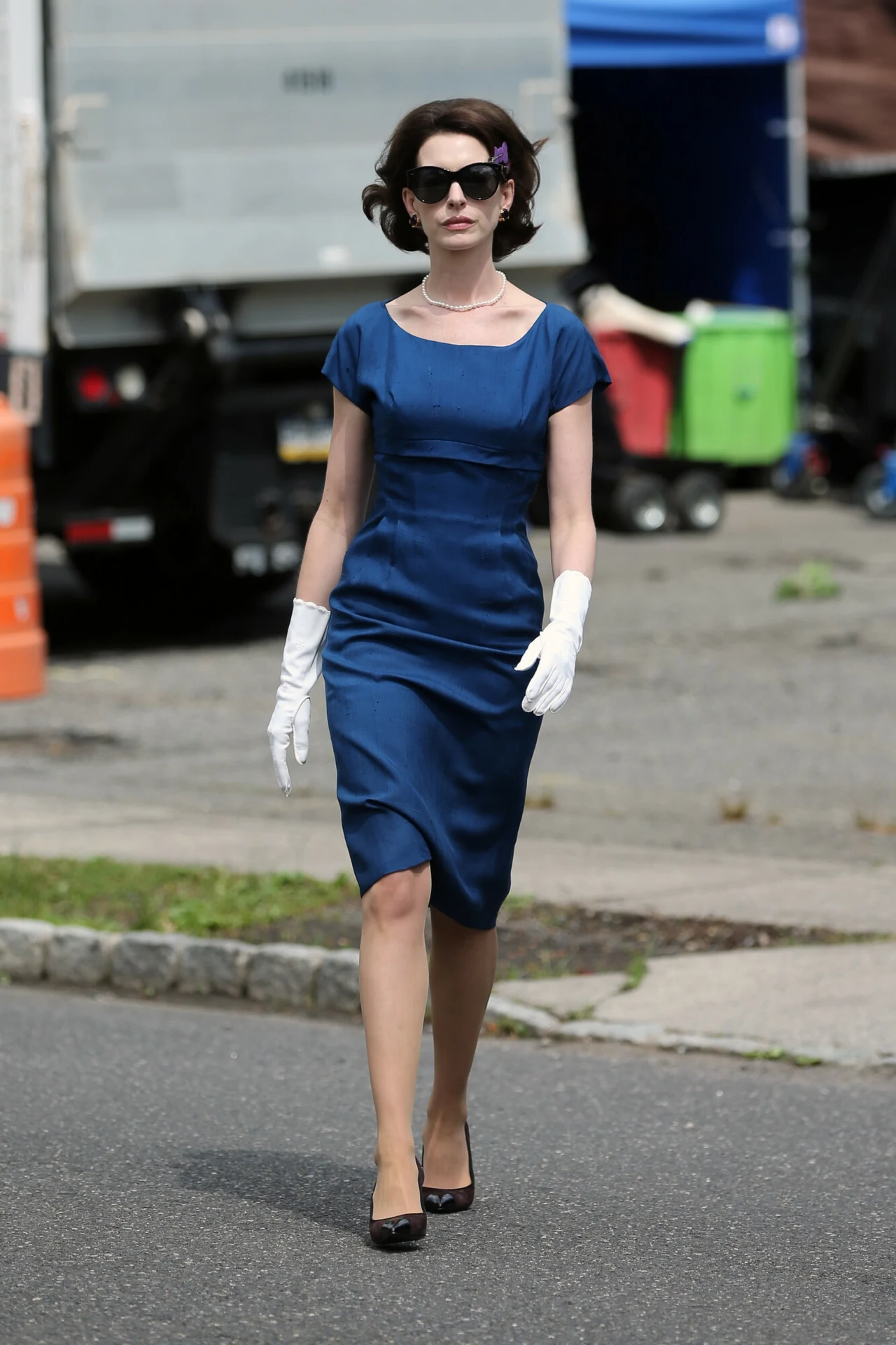 Kobieta ubrana w elegancką, niebieską sukienkę, białe rękawiczki i ciemne okulary przeciwsłoneczne idzie chodnikiem. W tle widoczne elementy miejskiego krajobrazu, takie jak samochód dostawczy, pojemniki na śmieci i fragmenty ulicy.