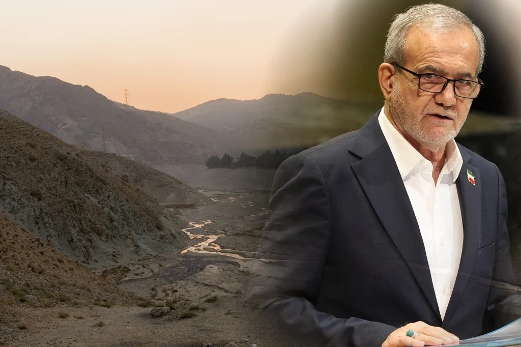 Starszy mężczyzna w okularach przemawia przy mównicy, nosi garnitur z białą koszulą i broszką na klapie. W tle widoczne góry o zmierzchu oraz suchy, kamienisty krajobraz z małą rzeką.