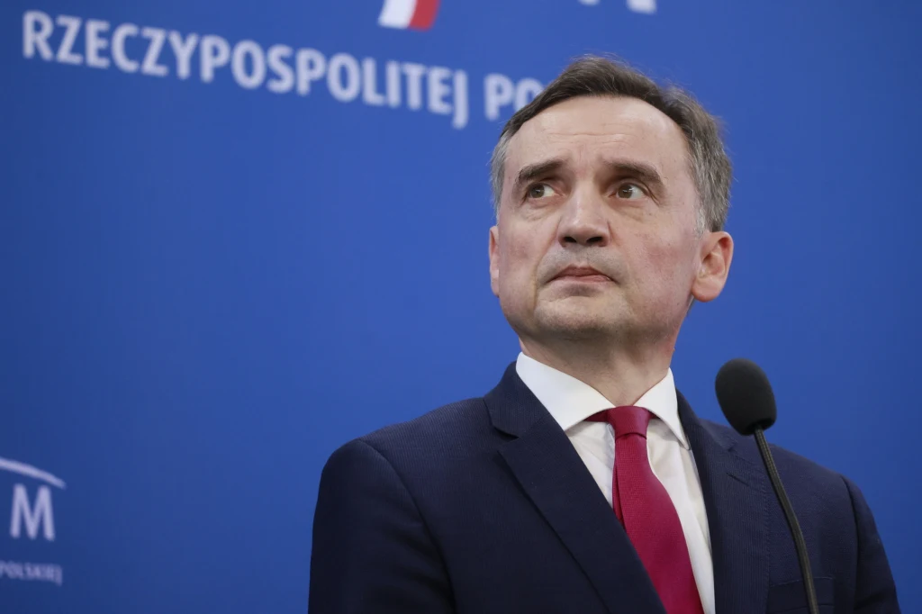 Mężczyzna w garniturze i czerwonym krawacie stoi przy mikrofonie na tle niebieskiej ściany z napisem Rzeczpospolitej Polskiej.