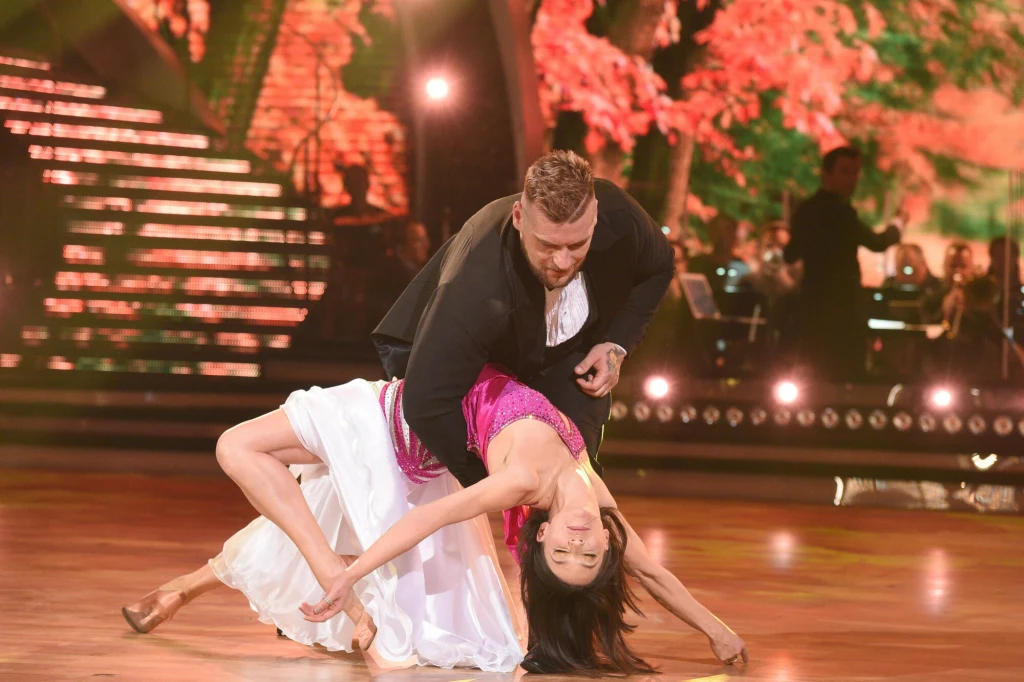 Para tancerzy wykonuje dynamiczną pozę taneczną na parkiecie, kobieta ubrana w białą spódnicę i różowy top jest mocno odchylona do tyłu, podtrzymywana przez partnera ubranego w czarny garnitur. W tle jasne światła i jesienne dekoracje wzmacniają teatra...