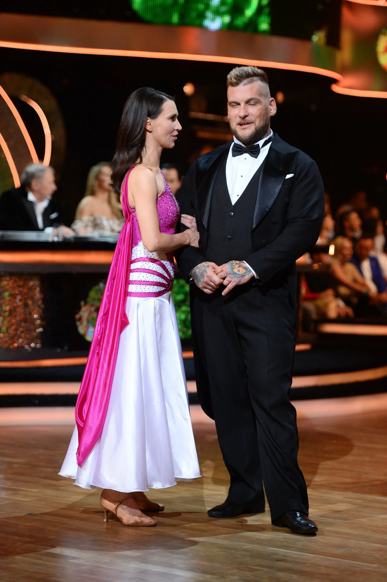 Para w eleganckich strojach tanecznych stoi na parkiecie, wokół widownia i jury programu telewizyjnego w tle, światła sceniczne podkreślają atmosferę wydarzenia.