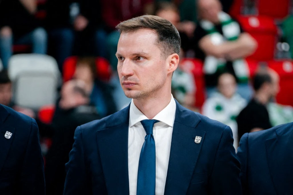 Mężczyzna w garniturze i krawacie na tle rozmytego tłumu na trybunach, skupiony i patrzący w bok.