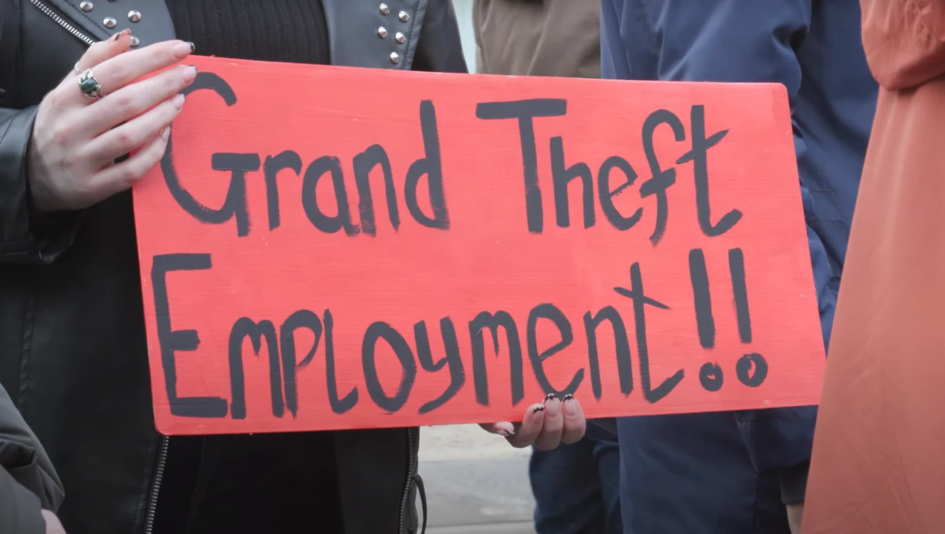 fragment filmu na kanale YouTube @People Make Games Ręka trzymająca czerwoną tabliczkę z napisem Grand Theft Employment!!, wokół widoczne osoby ubrane w ciemne ubrania.
