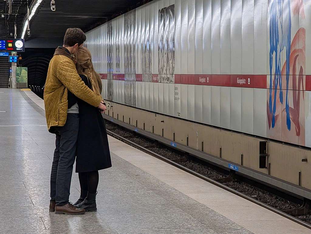 Para obejmująca się na peronie stacji metra, w tle pusty tor i zamknięte drzwi wagonów metra, ściana z oznaczeniami przystanków oraz rozmazane graffiti.