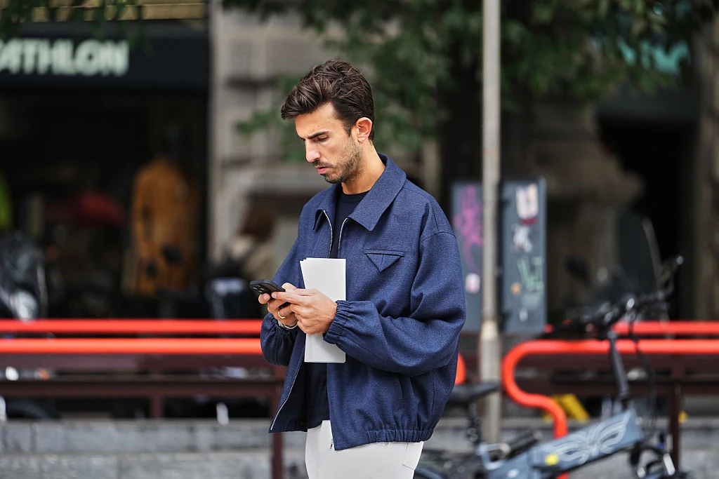 Mężczyzna w niebieskiej kurtce trzymający w jednej ręce biały dokument, a w drugiej smartfon, skoncentrowany na ekranie telefonu, w tle rozmyte elementy miejskiego krajobrazu i stojące rowery.