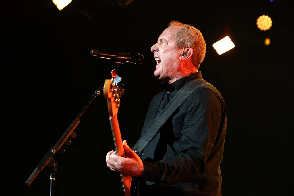 Andy McCluskey (Orchestral Manoeuvres in the Dark) w akcji Mężczyzna w ciemnej koszuli śpiewa do mikrofonu podczas koncertu, grając na gitarze basowej, podświetlony reflektorami scenicznymi.