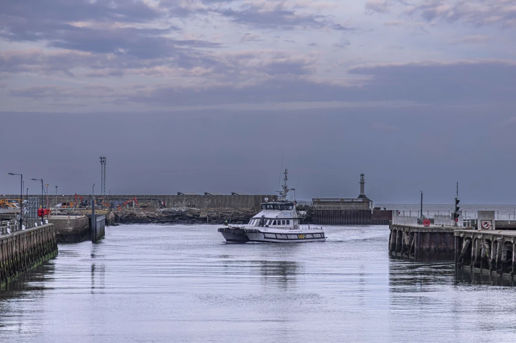 Statek w porcie otoczonym betonowymi nabrzeżami, spokojna woda i umiarkowanie zachmurzone niebo; w tle widać latarnię morską oraz zabudowania portowe.