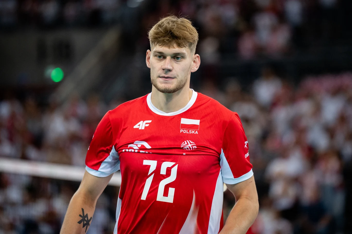 Siatkarz w czerwonej koszulce z numerem 12 i napisem Polska, stojący na hali sportowej, w tle rozmyte sylwetki kibiców.