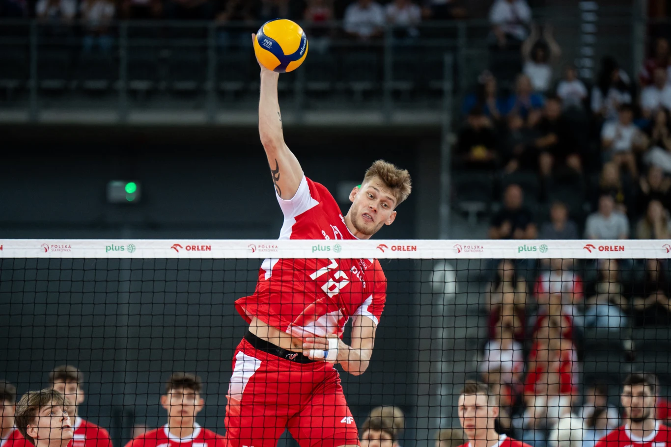 Zawodnik w czerwonym stroju siatkarskim wykonuje dynamiczny atak nad siatką podczas meczu, trzymając piłkę w uniesionej ręce, w tle widoczni są inni zawodnicy oraz publiczność na trybunach.