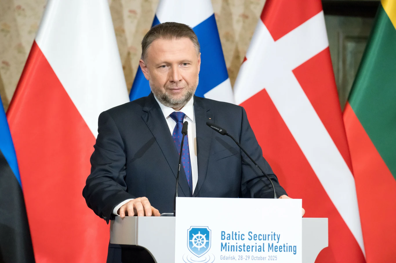 Marcin Kierwiński, szef MSWiA Mężczyzna w garniturze przemawiający przy mównicy, na tle flag państw bałtyckich oraz innych krajów, podczas spotkania ministrów poświęconego bezpieczeństwu w regionie Morza Bałtyckiego.