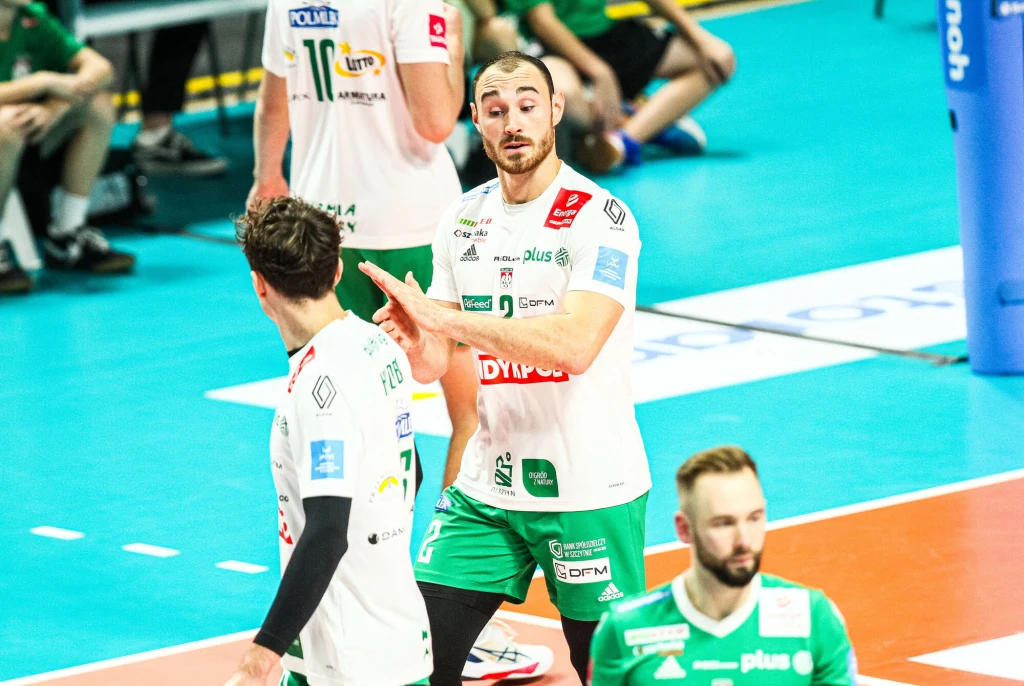 Moritz Karlitzek jest jednym z liderów Indykpolu AZS Olsztyn Zawodnicy drużyny siatkarskiej ubrani w białe i zielone stroje stoją na błękitnej hali sportowej podczas rozgrywki. Jeden z siatkarzy gestykuluje dłonią w kierunku kolegi z drużyny, w tle widoczni inni gracze oraz fragment boiska.