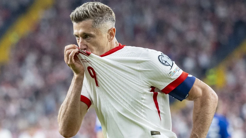 Robert Lewandowski Piłkarz ubrany w biały strój z numerem 9 całuje herb na koszulce, unosząc ją do ust, wyrażając emocje i przywiązanie do drużyny podczas meczu na tle rozmytego stadionu wypełnionego kibicami.