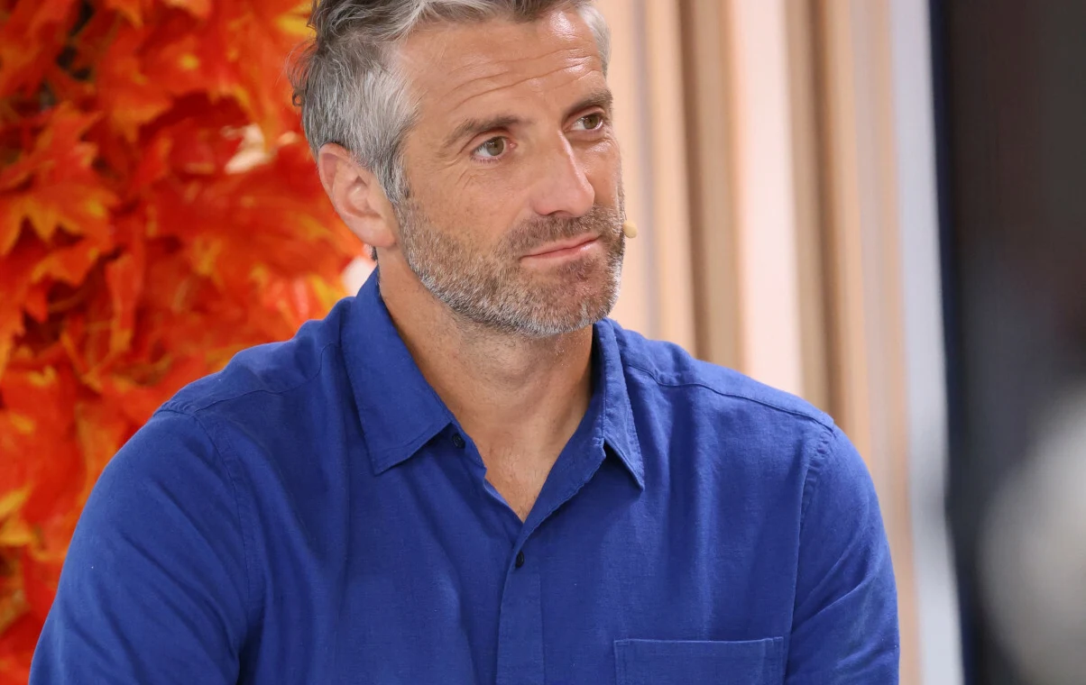 Maciej Dowbor Mężczyzna o siwych włosach i brodzie w niebieskiej koszuli siedzi na tle jesiennych liści oraz jasnych zasłon, patrząc lekko na bok.