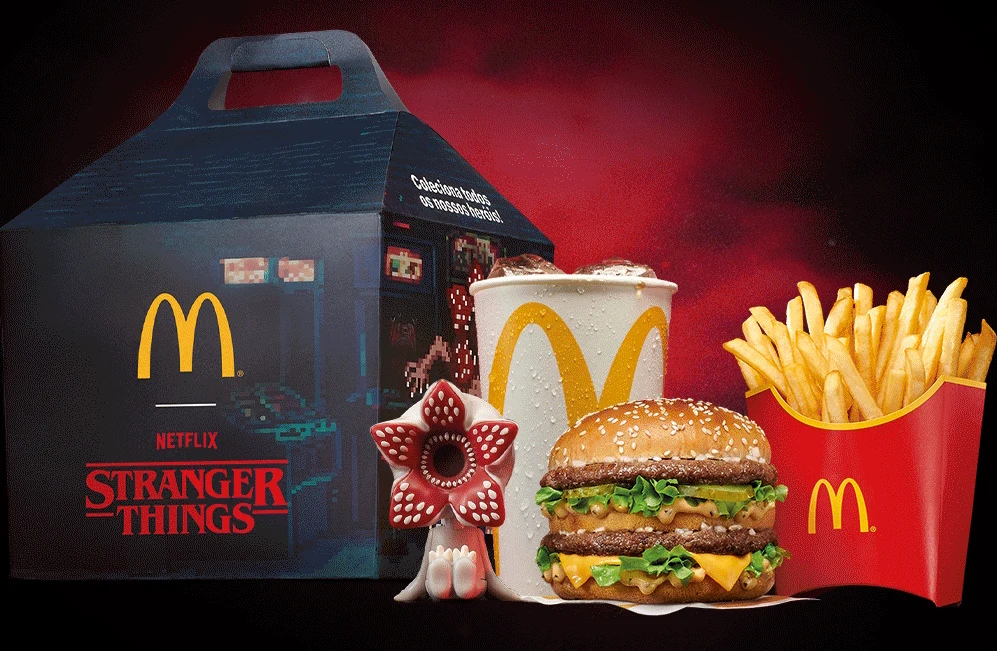 Zestaw jedzenia typu fast food z burgerem, frytkami i napojem, opakowanymi w pudełko z logo McDonald's oraz motywem serialu Stranger Things. W centrum znajduje się burger z dwoma kotletami i warzywami, obok pojemnik z frytkami oraz kubek z napojem. Dod...