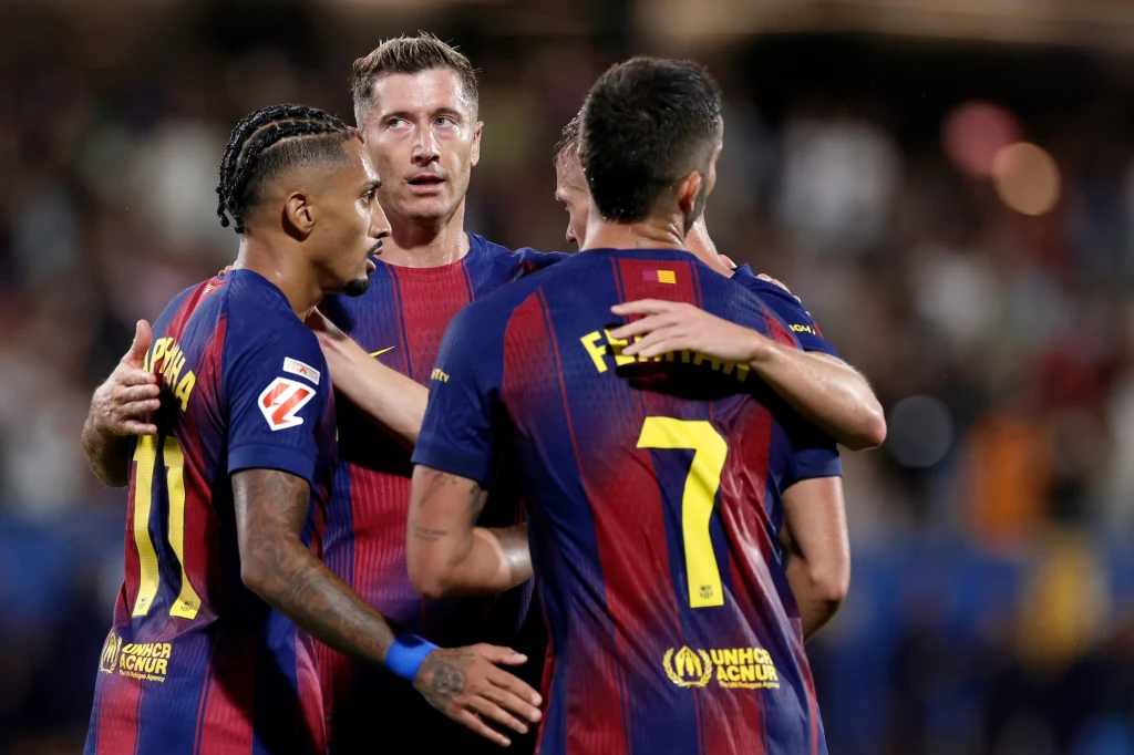 Grupa piłkarzy FC Barcelony w granatowo-bordowych strojach świętuje zdobycie gola, trzymając się w uścisku na murawie stadionu, wokół nich widać rozmytych kibiców w tle.