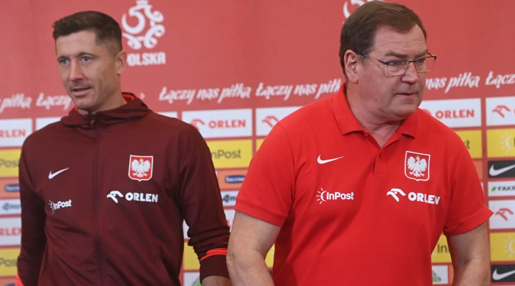 Robert Lewandowski w towarzystwie selekcjonera Jana Urbana Dwóch mężczyzn w sportowych ubraniach z emblematami polskiej reprezentacji na tle ściany z logotypami sponsorów i PZPN, skupieni podczas konferencji prasowej.