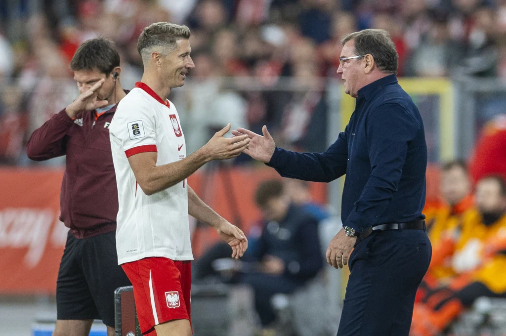 Robert Lewandowski i Jan Urban, 07.09.2025 r. Piłkarz w biało-czerwonym stroju reprezentacji Polski ściska dłoń trenera przy linii bocznej boiska. Obok nich mężczyzna z obsługi technicznej, a w tle rozmyta publiczność oraz ławka zawodników.