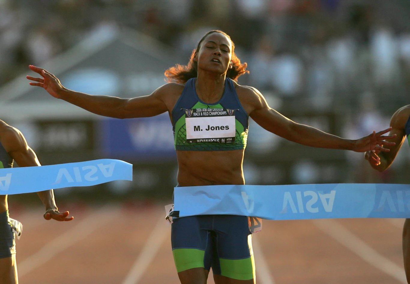 Lekkoatletka przekracza linię mety podczas wyścigu, mając rozłożone ramiona w geście zwycięstwa, z widocznym numerem startowym i tabliczką z nazwiskiem na piersi; w tle rozmyty stadion sportowy.