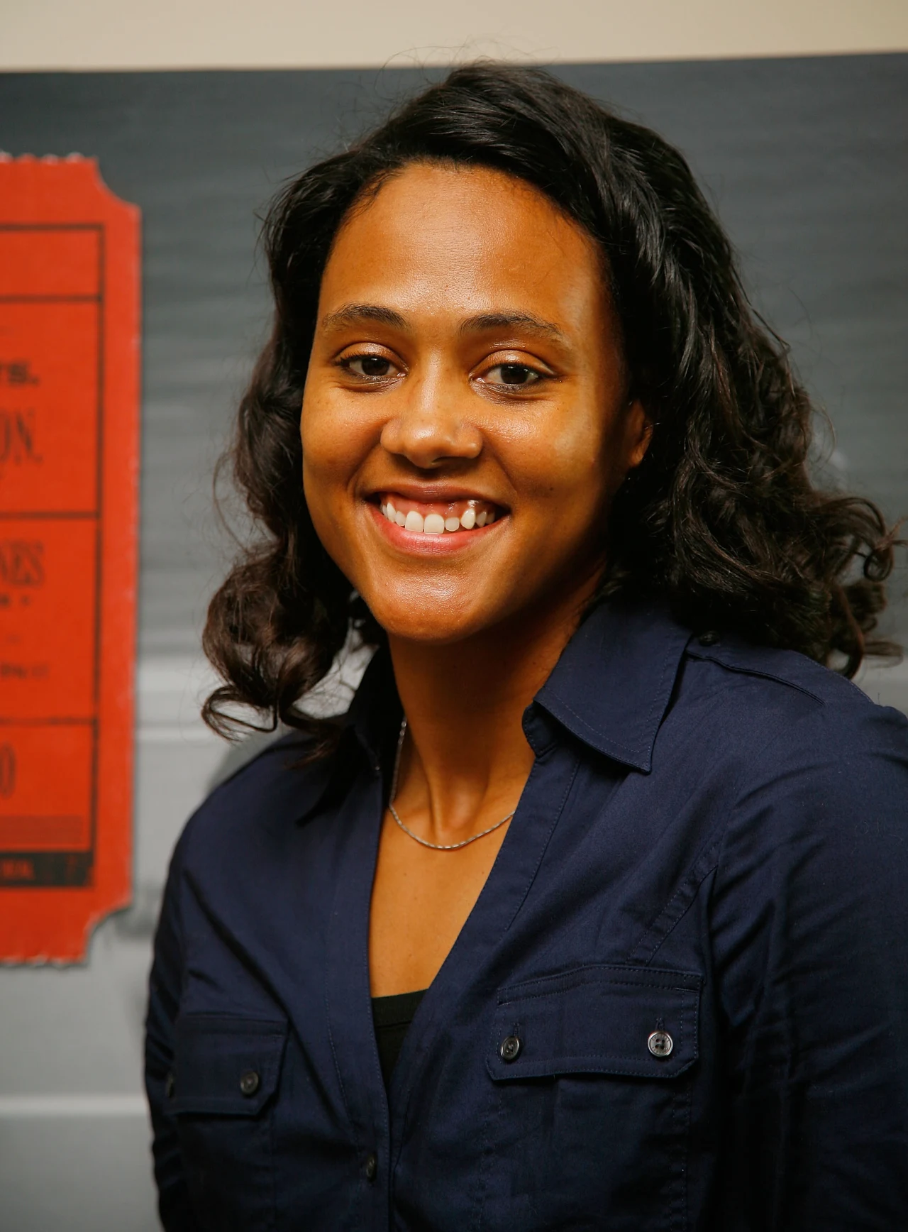 Uśmiechnięta kobieta o ciemnych włosach, ubrana w granatową koszulę, stoi na tle plakatu z czerwonym elementem po lewej stronie.