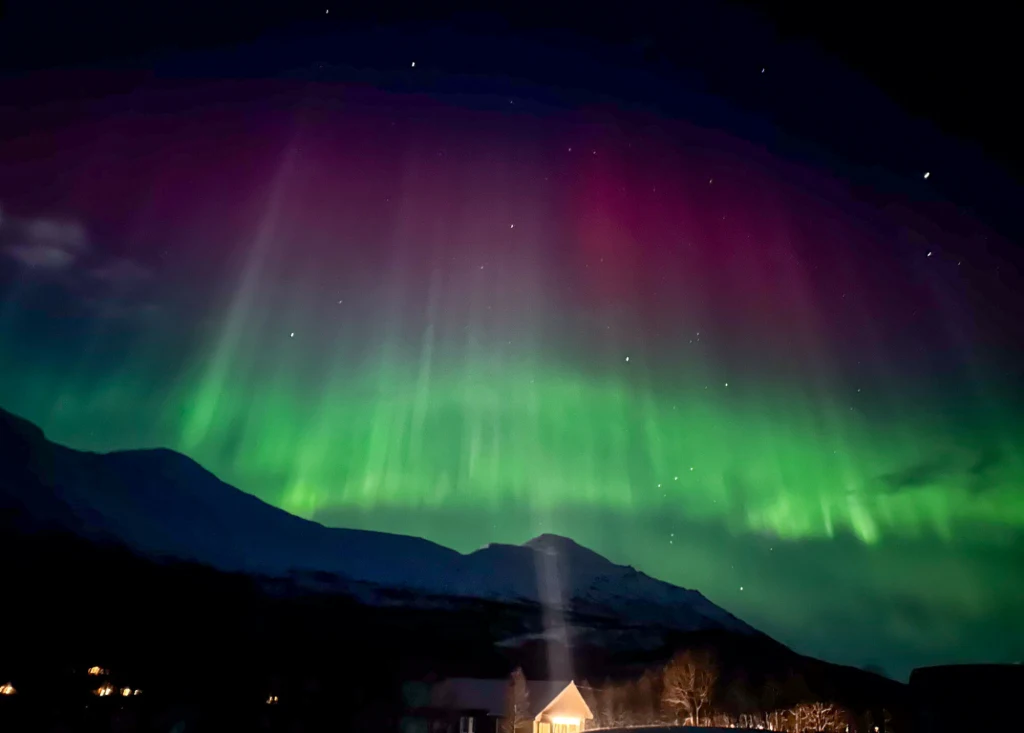Intensywna zorza polarna rozświetlająca nocne niebo barwami zieleni i fioletu nad górskim krajobrazem, z widocznym światłem domku w dolinie.
