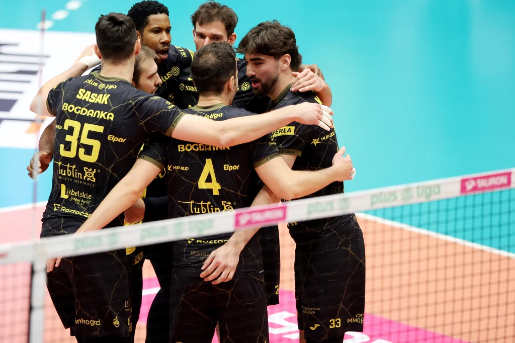 Siatkarze Bogdanki LUK Lublin Grupa zawodników drużyny siatkarskiej w czarnych strojach z żółtymi napisami świętuje zdobycie punktu przy siatce na parkiecie sportowej hali.