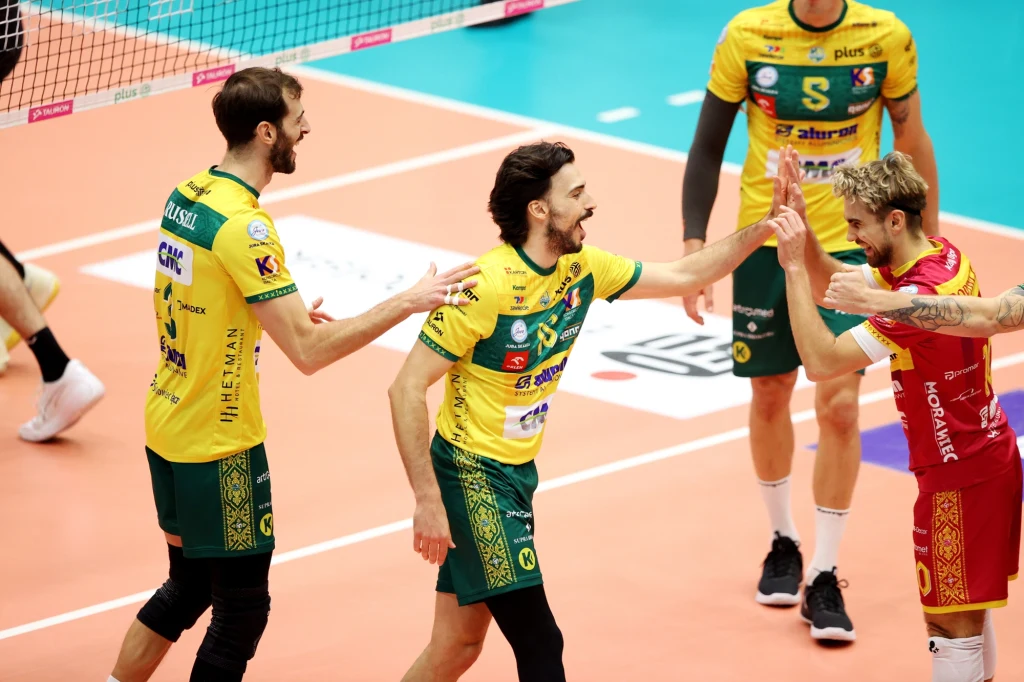 Siatkarze Aluron CMC Warty Zawiercie Trzech siatkarzy świętuje zdobycie punktu podczas meczu siatkówki, wszyscy ubrani w stroje sportowe, a dwóch z nich ubranych jest w zielono-żółte koszulki i spodenki, natomiast jeden w czerwono-żółtej odzieży. Jasne boisko i siatka siatkarska w tle.