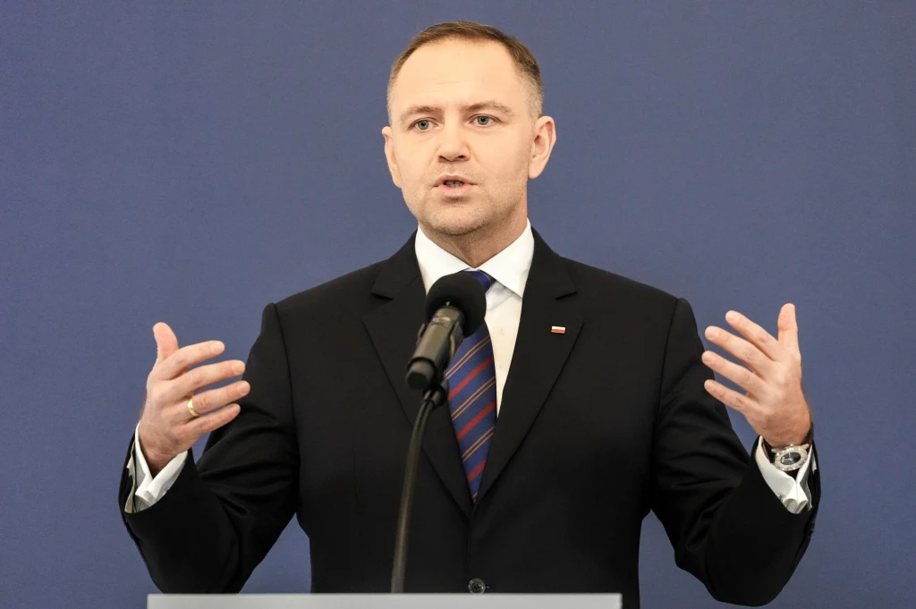 Mężczyzna w garniturze i krawacie stoi za mównicą i przemawia do mikrofonu, gestykulując rękami na tle jednolitego, niebieskiego tła.