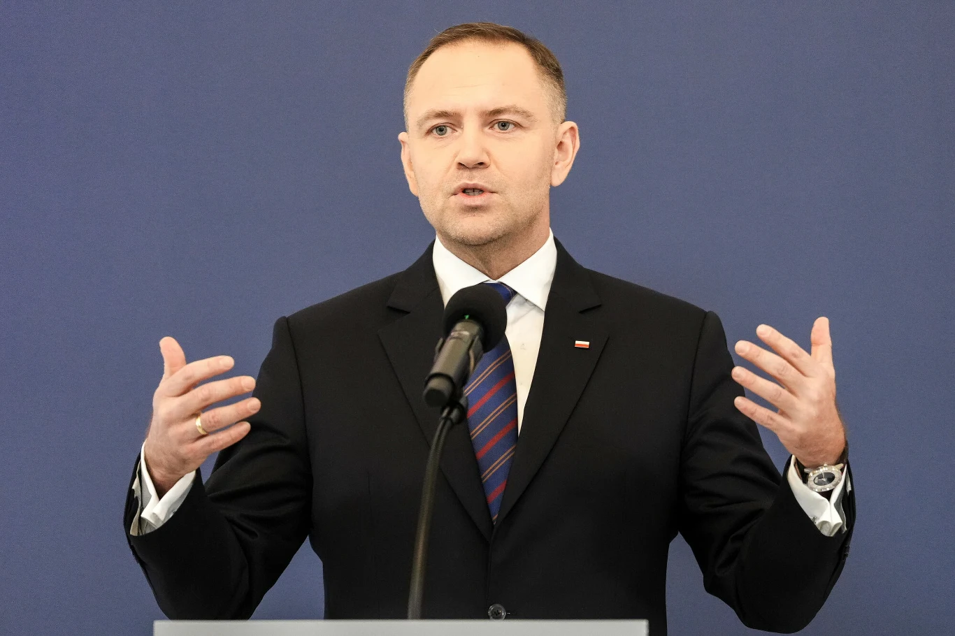 Karol Nawrocki zwrócił się z wnioskiem o delegalizację Komunistycznej Partii Polski Mężczyzna w garniturze i krawacie stoi za mównicą i przemawia do mikrofonu, gestykulując rękami na tle jednolitego, niebieskiego tła.