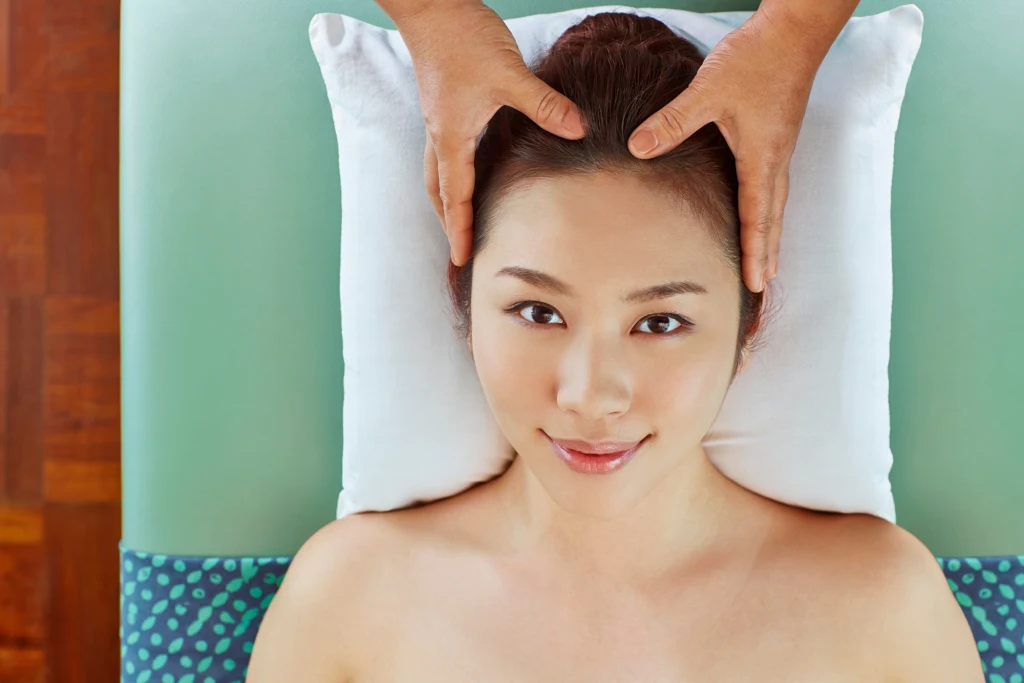 Jak dbać o włosy i skórę głowy? Kobieta leżąca na plecach zrelaksowana, z głową opartą na białej poduszce, poddawana masażowi głowy przez osobę stojącą poza kadrem.