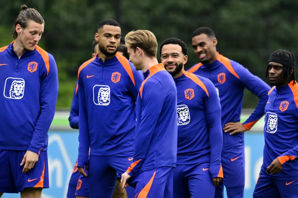 Grupa piłkarzy ubrana w niebiesko-pomarańczowe dresy reprezentacyjne stoi razem na boisku treningowym, zawodnicy są rozluźnieni i rozmawiają ze sobą, w tle widoczna jest zielona roślinność.