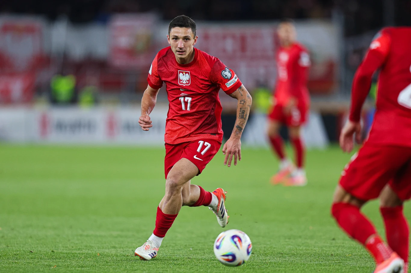 Bartosz Slisz Piłkarz w czerwonym stroju reprezentacji Polski biegnie z piłką po zielonej murawie podczas meczu piłkarskiego, widoczny z numerem 17 na koszulce. W tle inni zawodnicy oraz rozmyta publiczność na trybunach.